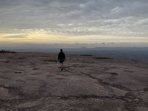 A person walks alone on a rocky landscape at sunset, with a cloudy sky and distant mountains in the background.