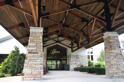 Entrance of a building featuring a wooden ceiling, stone pillars, and landscaped greenery.