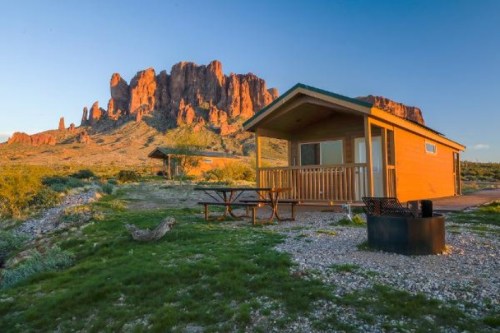 A cozy cabin with a porch sits in front of a rugged mountain landscape under a clear blue sky.