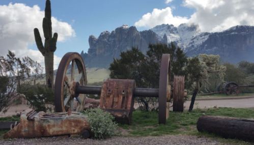 Rusty machinery in a desert landscape with mountains and snow-capped peaks in the background under a cloudy sky.