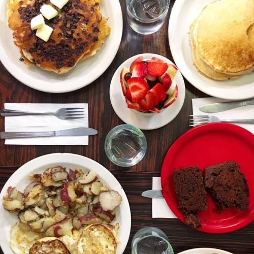 A table set with pancakes, chocolate chip pancakes, fruit, potatoes, and a slice of chocolate cake.