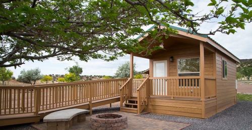 A wooden cabin with a ramp, surrounded by trees and a stone fire pit, set in a spacious outdoor area.