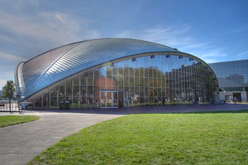 A modern glass building with a curved roof, surrounded by green grass and trees under a clear blue sky.