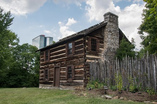 A rustic log cabin with a stone chimney, surrounded by a wooden fence, set against a backdrop of modern buildings.