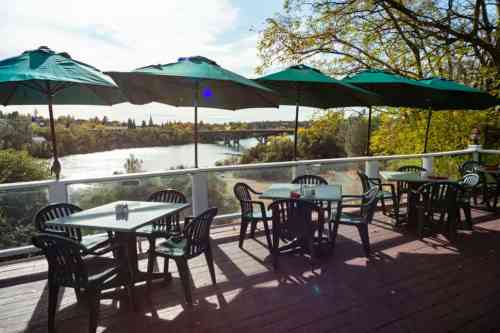 Outdoor dining area with green umbrellas overlooking a river and trees under a bright sky.