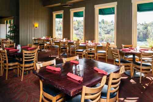 Cozy restaurant interior with wooden tables, chairs, and large windows showcasing a scenic view outside.