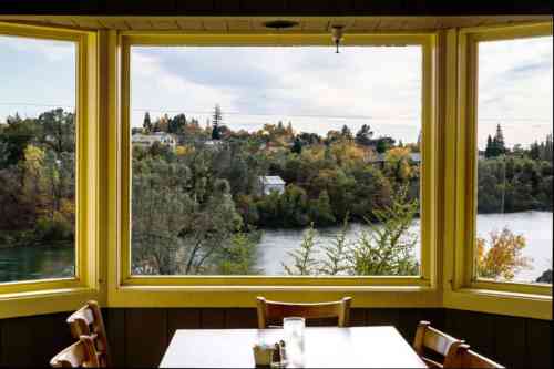 A scenic view of a river and trees through large yellow-framed windows, with a table set for dining in the foreground.