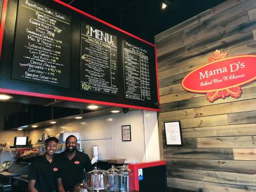 A restaurant interior featuring a menu board and two staff members smiling behind the counter at Mama D's.