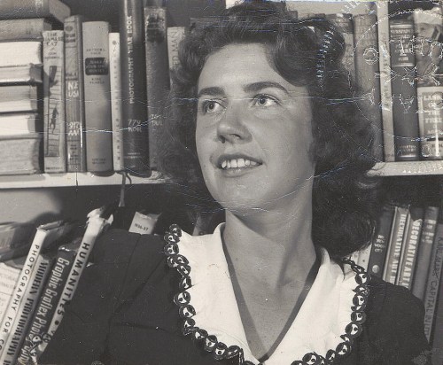 A smiling woman with curly hair, wearing a black dress with a white collar, stands in front of a bookshelf filled with books.