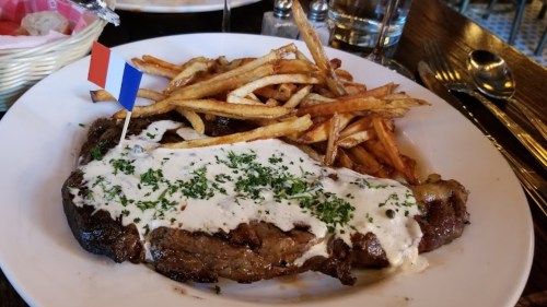 A plate of steak topped with creamy sauce and herbs, served with crispy French fries and a small French flag.