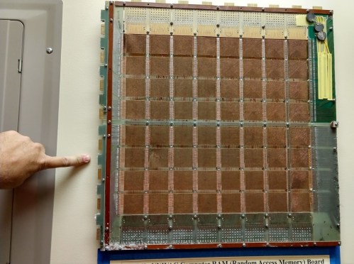 A hand points at a large, vintage RAM board with a grid of copper and metal components mounted on a green base.
