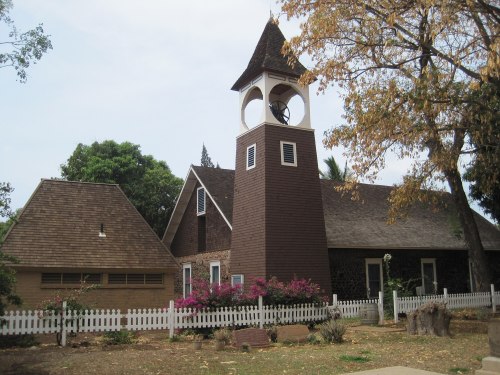 A brown church with a tall bell tower, surrounded by trees and a white picket fence, featuring blooming flowers in front.
