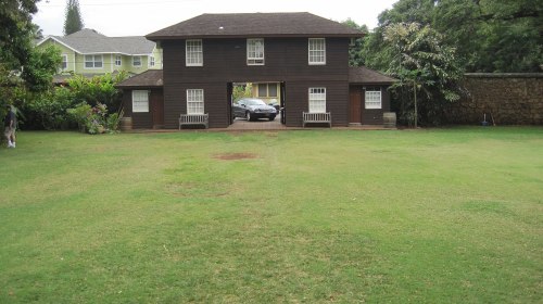 A two-story brown house with a car parked in front, surrounded by a green lawn and trees.