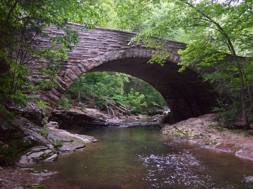 A stone arch bridge spans a serene creek, surrounded by lush green trees and rocky banks.