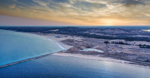 Aerial view of a serene beach at sunset, with calm waters and a sandy shoreline under a colorful sky.