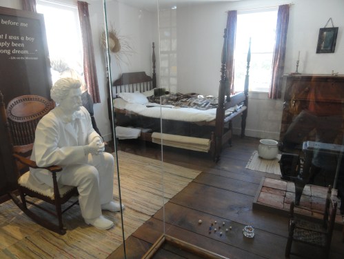 A glass-enclosed room featuring a life-sized statue of a boy in a white suit, a bed, and rustic furnishings.