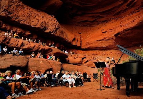 A musician performs in a red rock amphitheater, with an audience seated on the rocks and chairs.