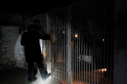 A person in dark clothing shines a flashlight on a barred prison cell door in a dimly lit space.