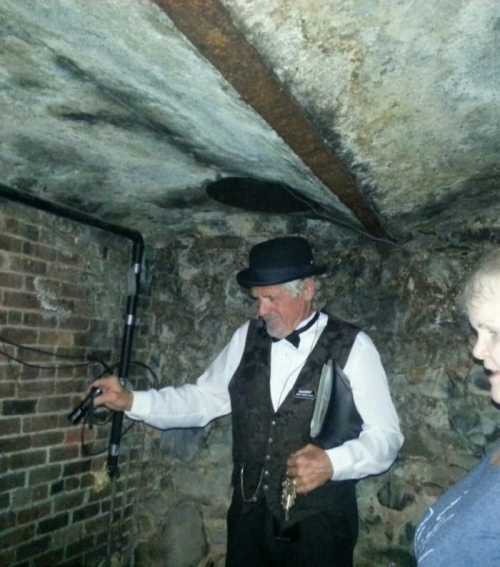 A man in a suit and hat stands in a dimly lit, brick-walled space, holding a device and speaking to another person.