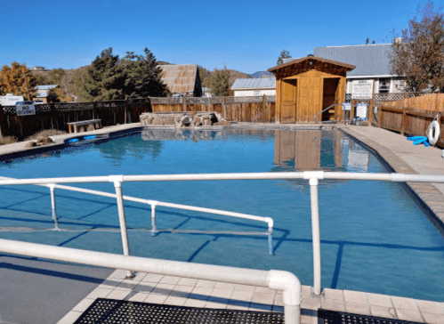 A clear swimming pool surrounded by a wooden fence, with a small shed and trees in the background under a blue sky.