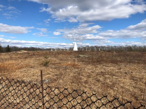 A grassy field with a white monument in the distance, under a blue sky with scattered clouds, bordered by a chain-link fence.
