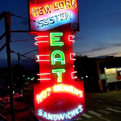 Bright neon sign reading "EAT HOT WEINERS SANDWICHES" against a twilight sky, with "NEW YORK SYSTEM" above.