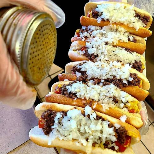 A hand sprinkles onions on a row of hot dogs topped with chili and mustard, arranged on a counter.
