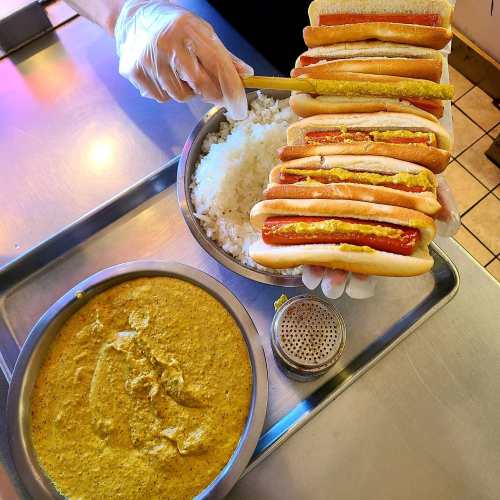 A person in gloves holds a stack of hot dogs with mustard over a bowl of rice and a tray of sauce.