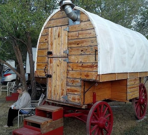 A wooden wagon with a white canopy, featuring red wheels, parked under trees, with a person sitting nearby.