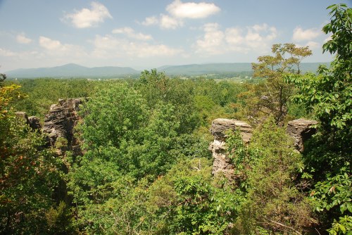 Lush green trees surround rocky cliffs, with distant mountains under a bright blue sky and fluffy white clouds.