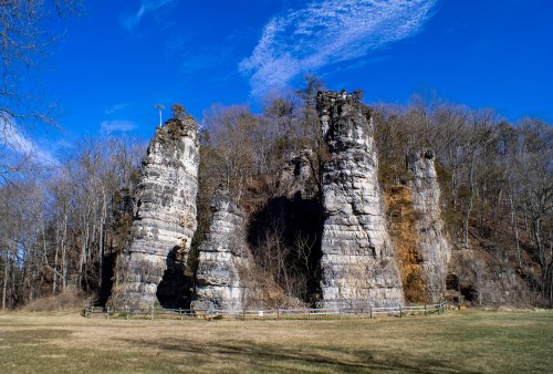 Tall rock formations rise against a clear blue sky, surrounded by trees and a grassy area.