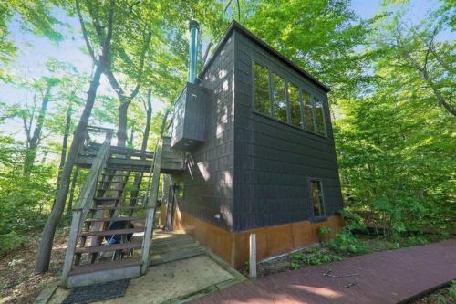 A modern black cabin surrounded by lush green trees, featuring a staircase and large windows.