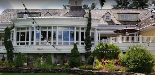 A large, elegant house with a curved porch, surrounded by greenery and decorative string lights.