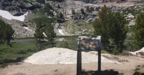 A scenic view of a lake surrounded by rocky terrain and trees, with an informational sign in the foreground.