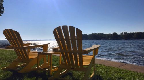 Two yellow Adirondack chairs facing a calm lake under a clear blue sky, with gentle waves lapping at the shore.