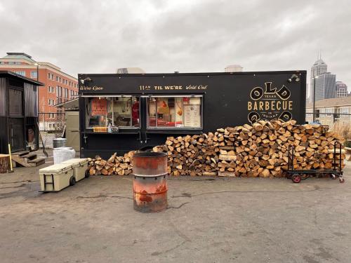 A food truck labeled "Old Texas Barbecue" with a stack of firewood in front, set against a cloudy sky.