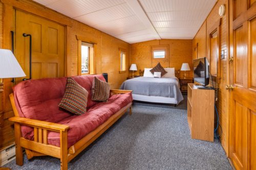 Cozy cabin interior featuring a bed, futon, TV, and wooden walls with warm lighting.