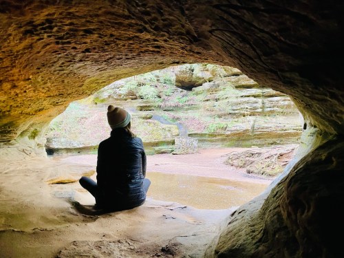 A person sits in a cave, looking out at a serene landscape with greenery and a small stream.