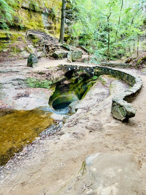 A stone bridge over a small stream in a lush, green forest with rocky terrain and moss-covered surfaces.