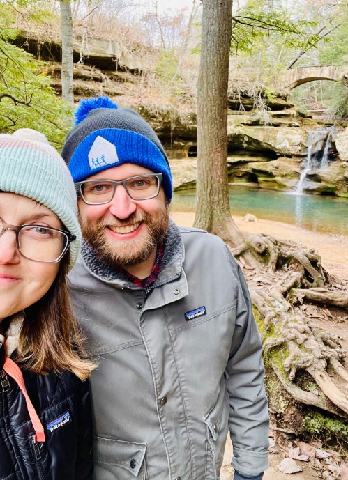 A smiling couple poses in front of a scenic forest with a waterfall and rocky landscape. Both wear winter hats.