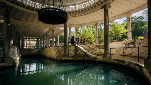 A serene indoor space with a circular pool, surrounded by columns and greenery, featuring a large chandelier above.