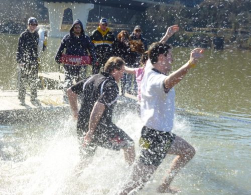 Two men running into a river, splashing water, while a group of people watches from the shore.