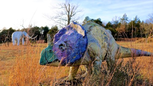A colorful dinosaur sculpture in a grassy field, with a white mammoth statue in the background under a clear sky.