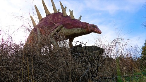 A colorful dinosaur sculpture with spines, partially hidden in tall grass against a blue sky.