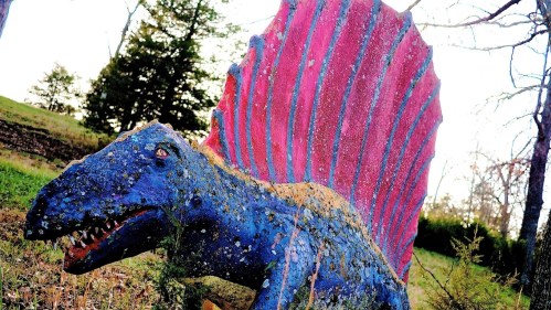 A colorful dinosaur sculpture with a spiked back, set in a grassy area surrounded by trees.