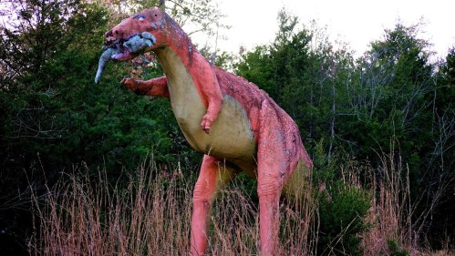 A large, colorful dinosaur statue stands in tall grass, holding a fish in its mouth, surrounded by trees.