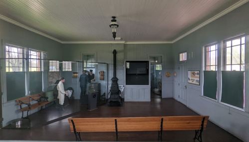 Interior of a historical building with benches, a stove, and displays, featuring two figures in period clothing.