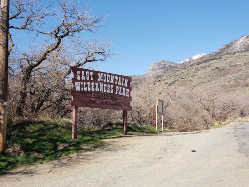 Sign for East Mountain Wilderness Park, surrounded by trees and mountains, with a dirt road leading into the park.
