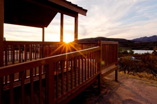 Sunset view from a wooden deck, with mountains and a lake in the background, casting warm light across the scene.