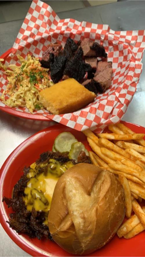 A plate of barbecue with brisket, coleslaw, cornbread, and fries, alongside a cheeseburger with pickles.